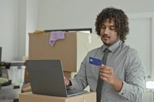 man holding creditcard and typing on laptop keyboard next to moving boxes in building; moving setup confidence